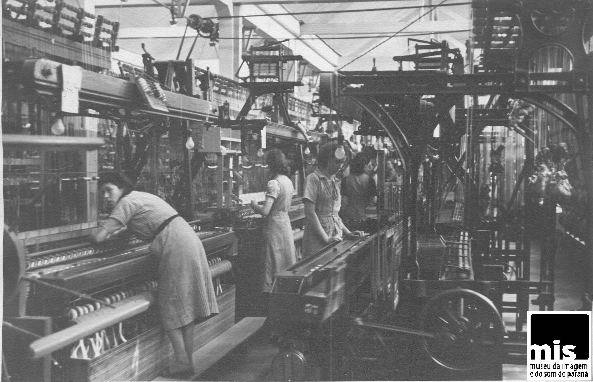 Descrição da imagem: fotografia horizontal em preto e branco. Imagem mostra interior do galpão de uma fábrica, com grandes teares mecânicos de produção de fitas, dispostos em fileiras. Nos corredores entre os teares, mulheres manuseiam as máquinas. As operárias usam uniforme composto de vestido com comprimento pouco abaixo do joelho e manga curta.  