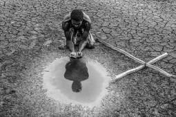 Homem pega água de uma poça com as mãos em terreno árido com uma cruz de madeira ao lado direito