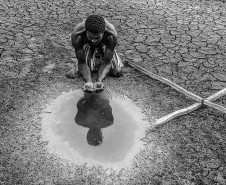 Homem pega água de uma poça com as mãos em terreno árido com uma cruz de madeira ao lado direito