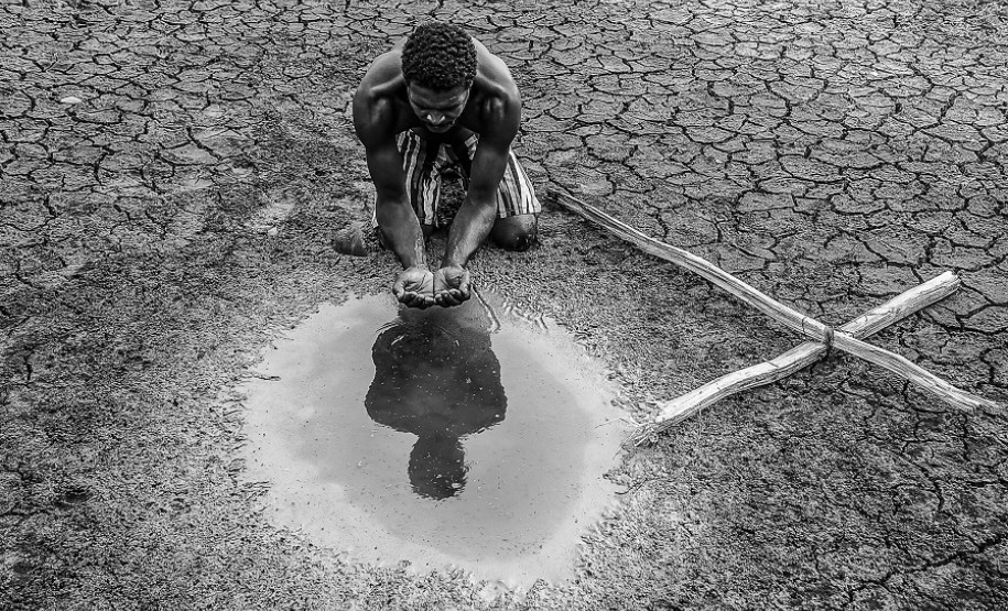 Homem pega água de uma poça com as mãos em terreno árido com uma cruz de madeira ao lado direito
